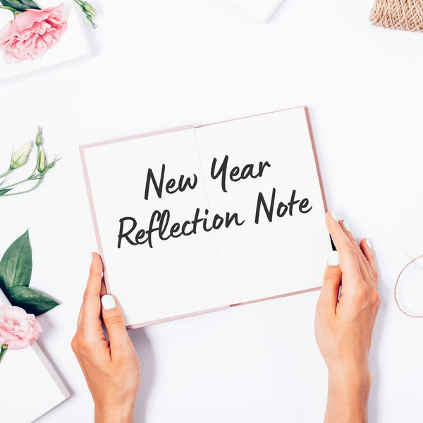 Hands holding a notebook titled 'New Year Reflection Note' on a white surface with flowers.